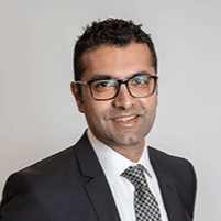 Alim Dhanji headshot. He is standing with hands in pockets wearing a dark suit and a checker board tie